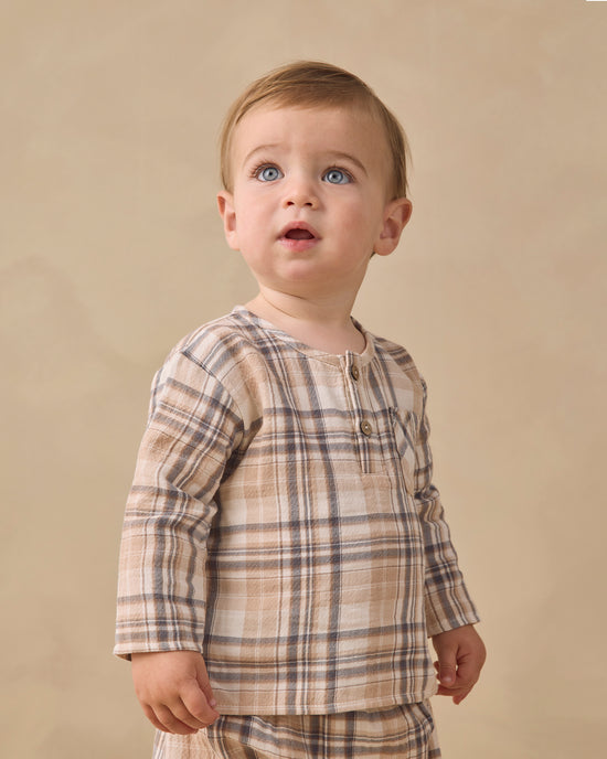 A baby boy wearing a beige and brown plaid long-sleeve henley shirt, standing against a solid beige studio background.