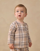 A baby boy wearing a beige and brown plaid long-sleeve henley shirt, standing against a solid beige studio background.