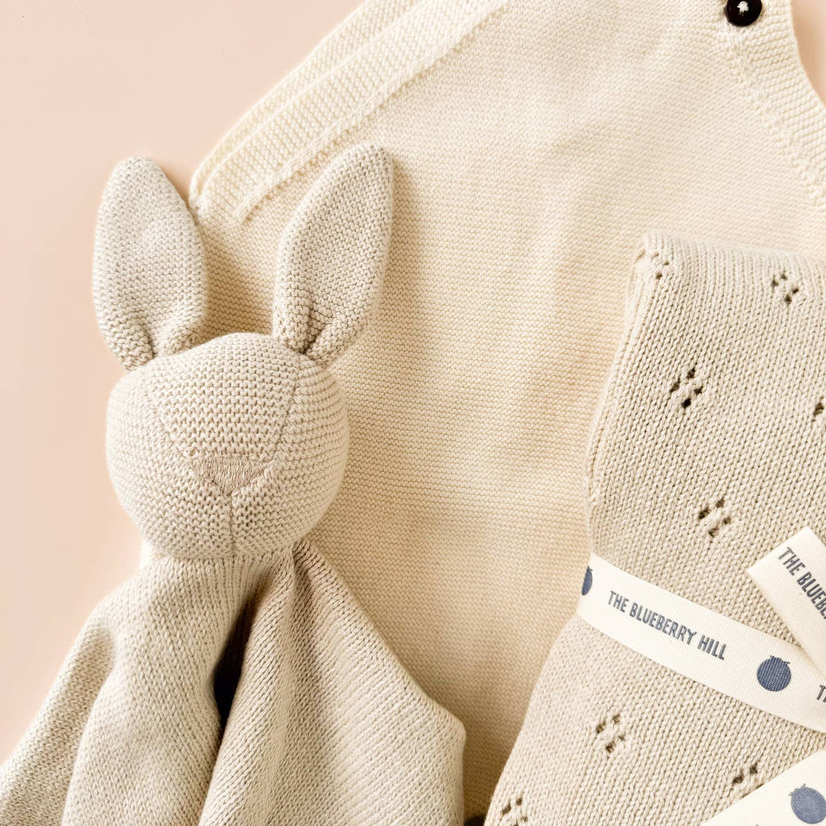Knitted baby blanket with bunny ears and 'The Blueberry Hill' label on a pink background