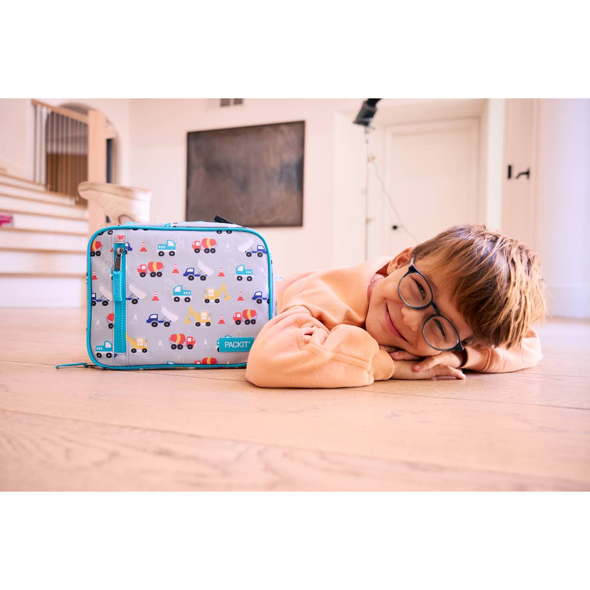 Child lying on a wooden floor next to a colorful suitcase with car patterns