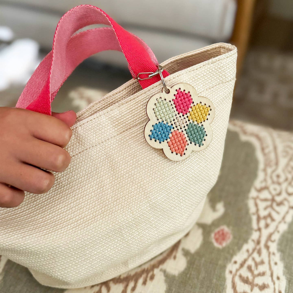 Beige straw bag with pink handle held by a hand, featuring a colorful flower-shaped tag.
