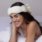 Woman wearing a white fleece headband with bow, sitting on a bed.