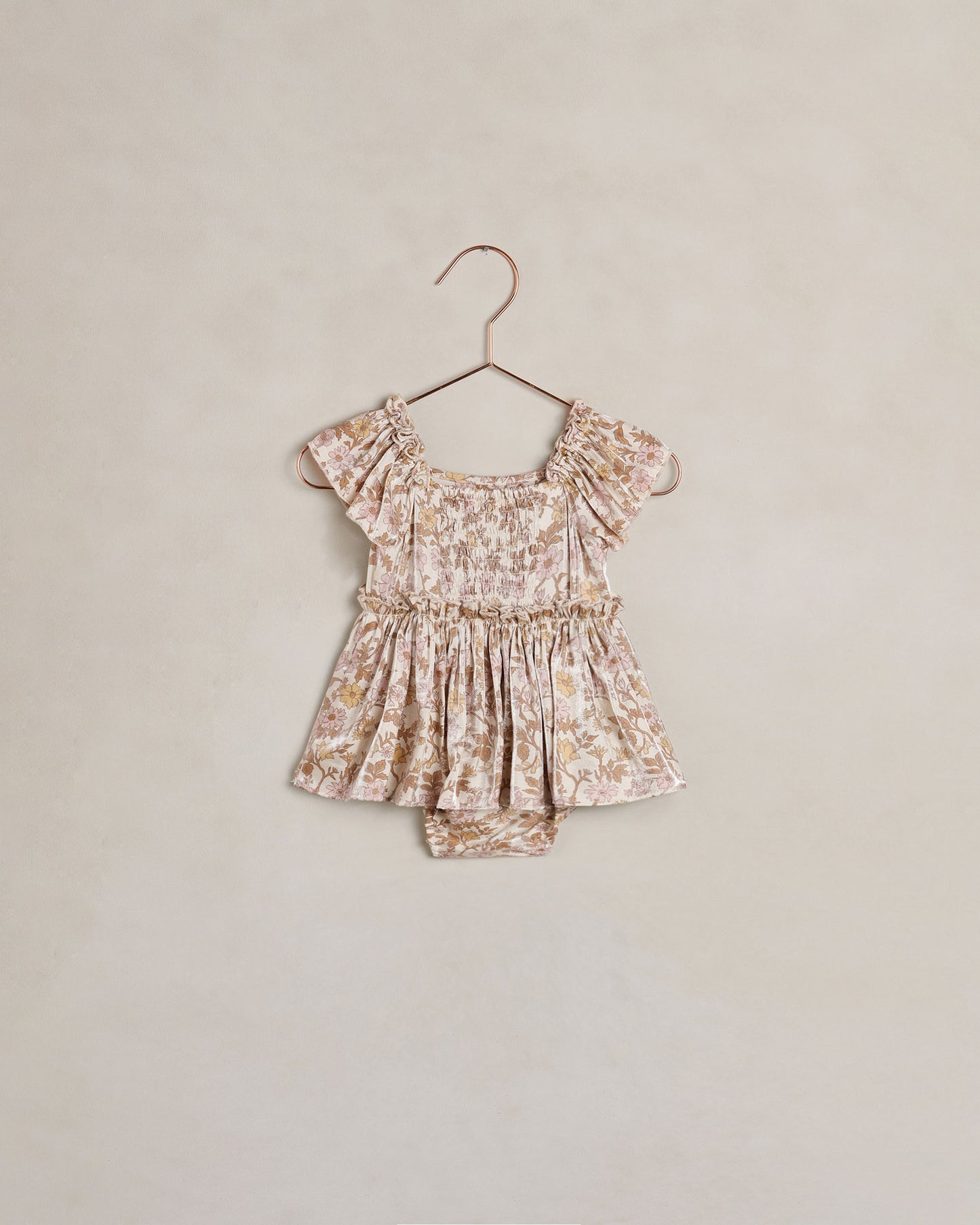 A toddler girl's short-sleeve romper in a beige and brown floral print, hanging on a metal hanger against a textured beige wall.
