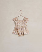 A toddler girl's short-sleeve romper in a beige and brown floral print, hanging on a metal hanger against a textured beige wall.