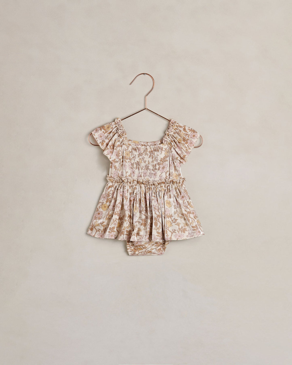 A toddler girl's short-sleeve romper in a beige and brown floral print, hanging on a metal hanger against a textured beige wall.