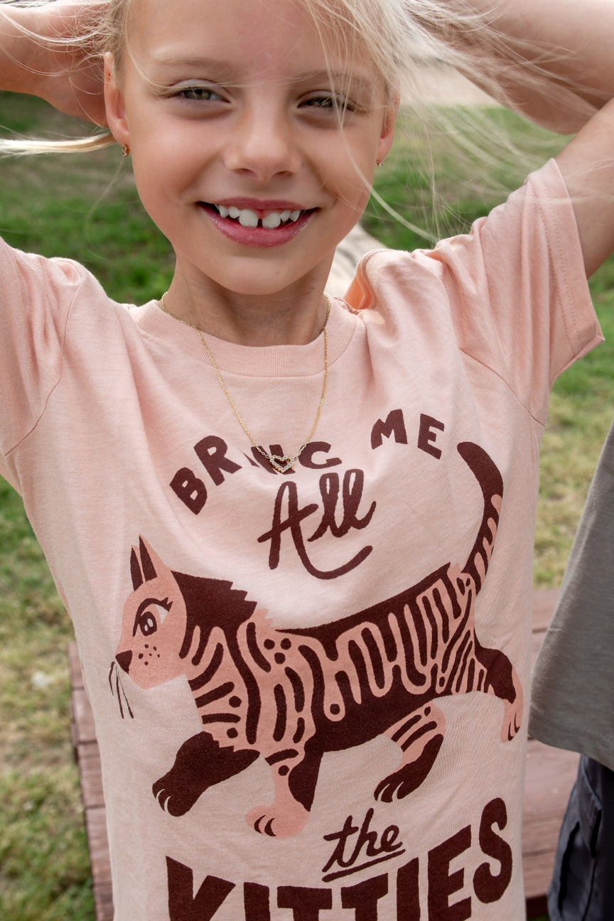 A young girl smiling outdoors while wearing a light pink graphic t-shirt with a dark brown cat design and the text 'BRING ME ALL THE KITTIES'.