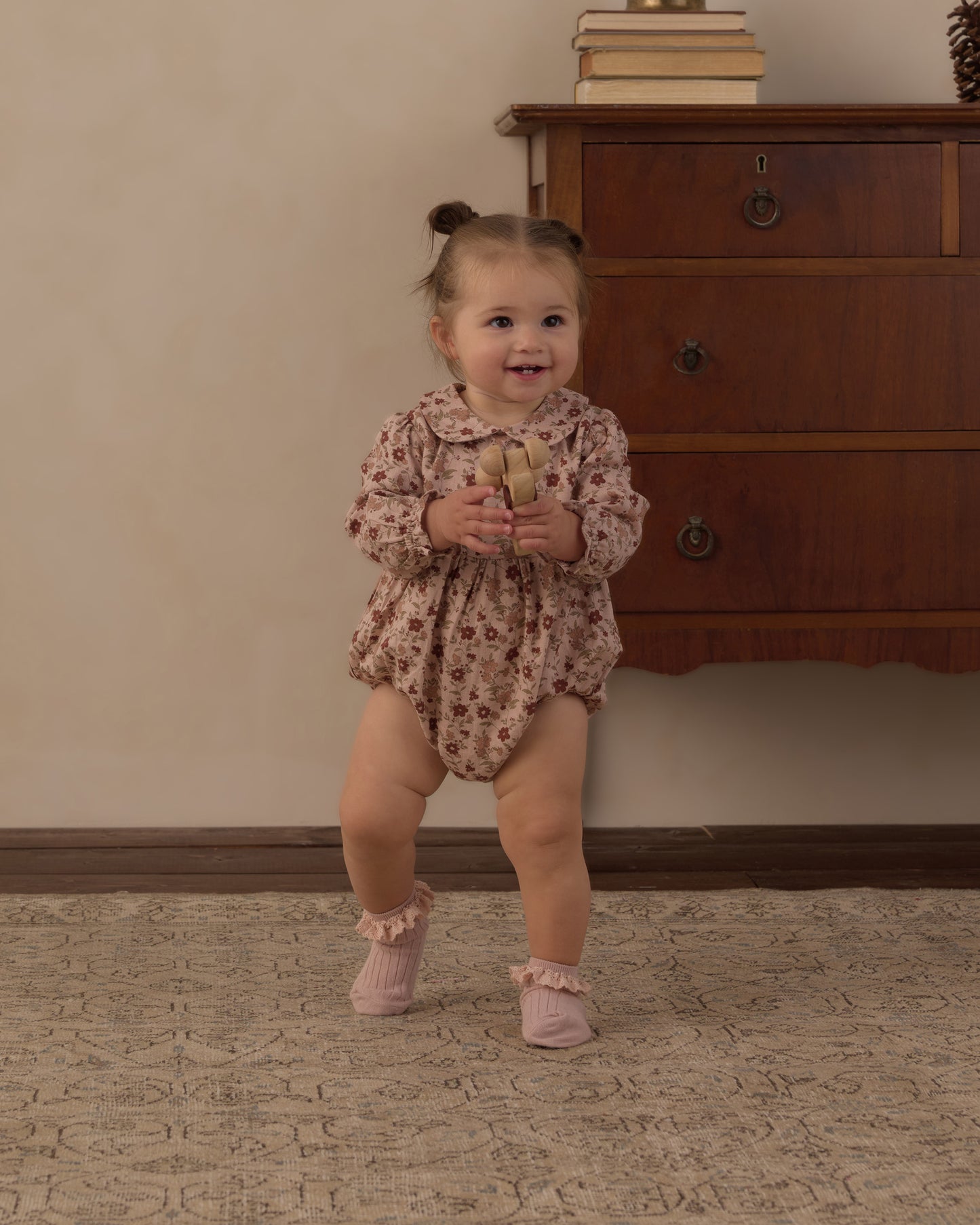 A toddler girl wearing a long-sleeve floral romper and pink booties stands on a patterned rug in front of a wooden dresser.