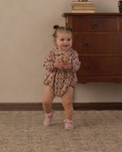 A toddler girl wearing a long-sleeve floral romper and pink booties stands on a patterned rug in front of a wooden dresser.