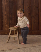 A toddler wearing Rylee + Cru Ruffle Collar Bodysuit in Natural Ditsy paired with Midnight ribbed leggings and a matching hair bow.