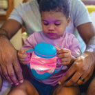 Child holding a pink and blue sippy cup with an adult's hands on either side.