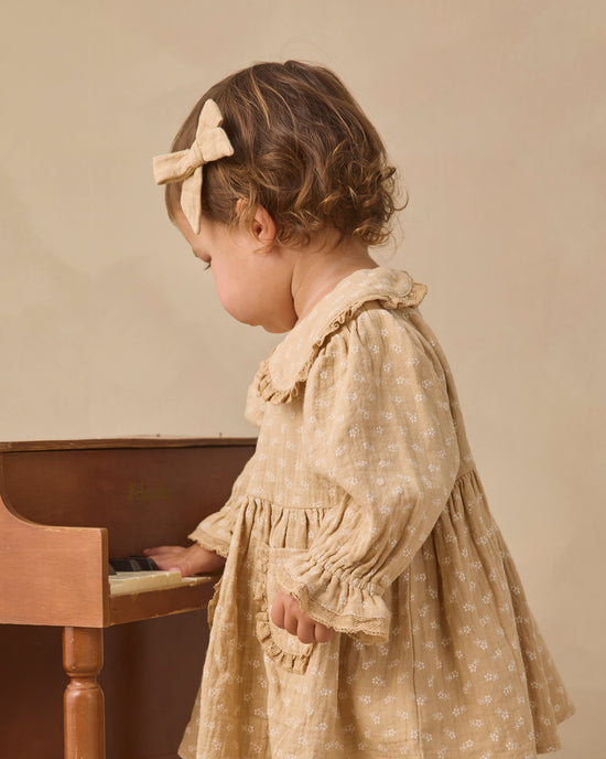 A toddler in profile wearing a beige floral dress and matching bow headband, standing beside a wooden piano against a warm beige background.