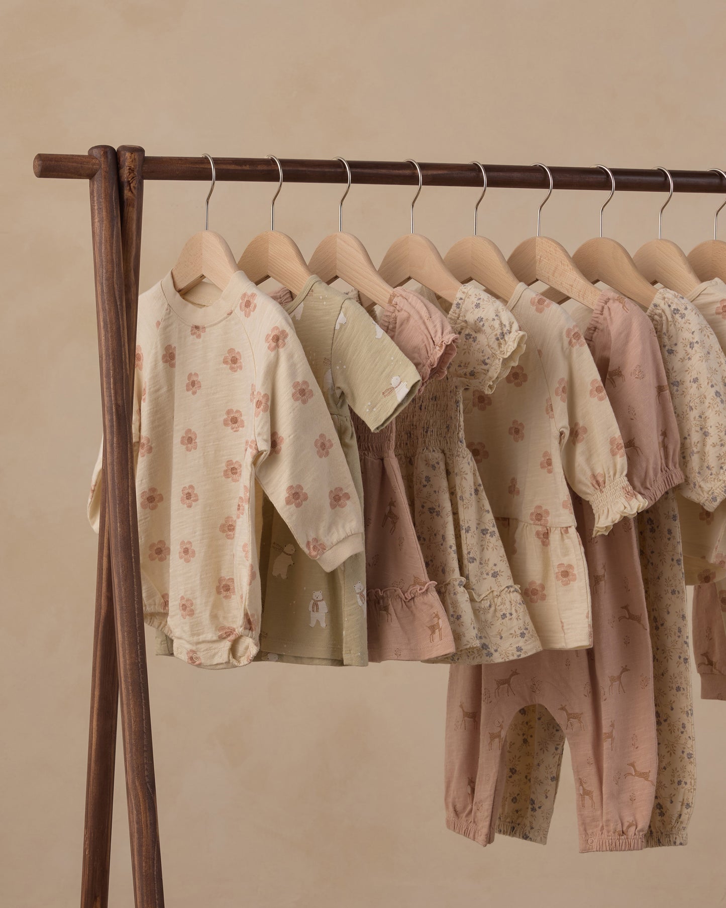 A wooden clothing rack holding multiple soft pastel baby outfits, including long-sleeve tops and pants, in cream, blush, and dusty pink tones with floral and animal prints, against a warm beige wall.