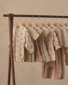 A wooden clothing rack holding multiple soft pastel baby outfits, including long-sleeve tops and pants, in cream, blush, and dusty pink tones with floral and animal prints, against a warm beige wall.