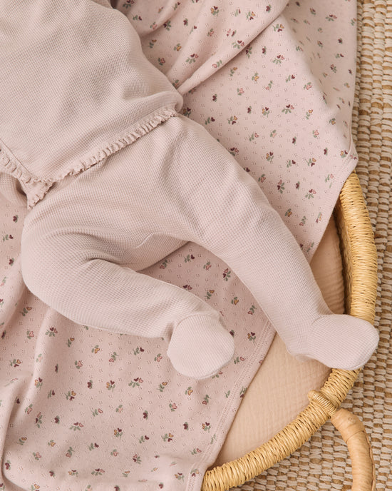 A close-up of a baby's legs wearing a pale pink waffle-knit footed sleeper, lying on a matching light pink floral blanket inside a woven rattan bassinet.