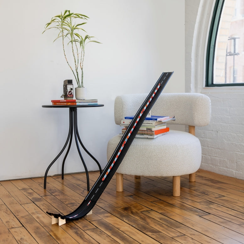 A black toy race track with red and white markings is set up on a wooden floor in a bright room, looping from near the bottom left toward a white armchair beside a small black side table with a potted plant and books.
