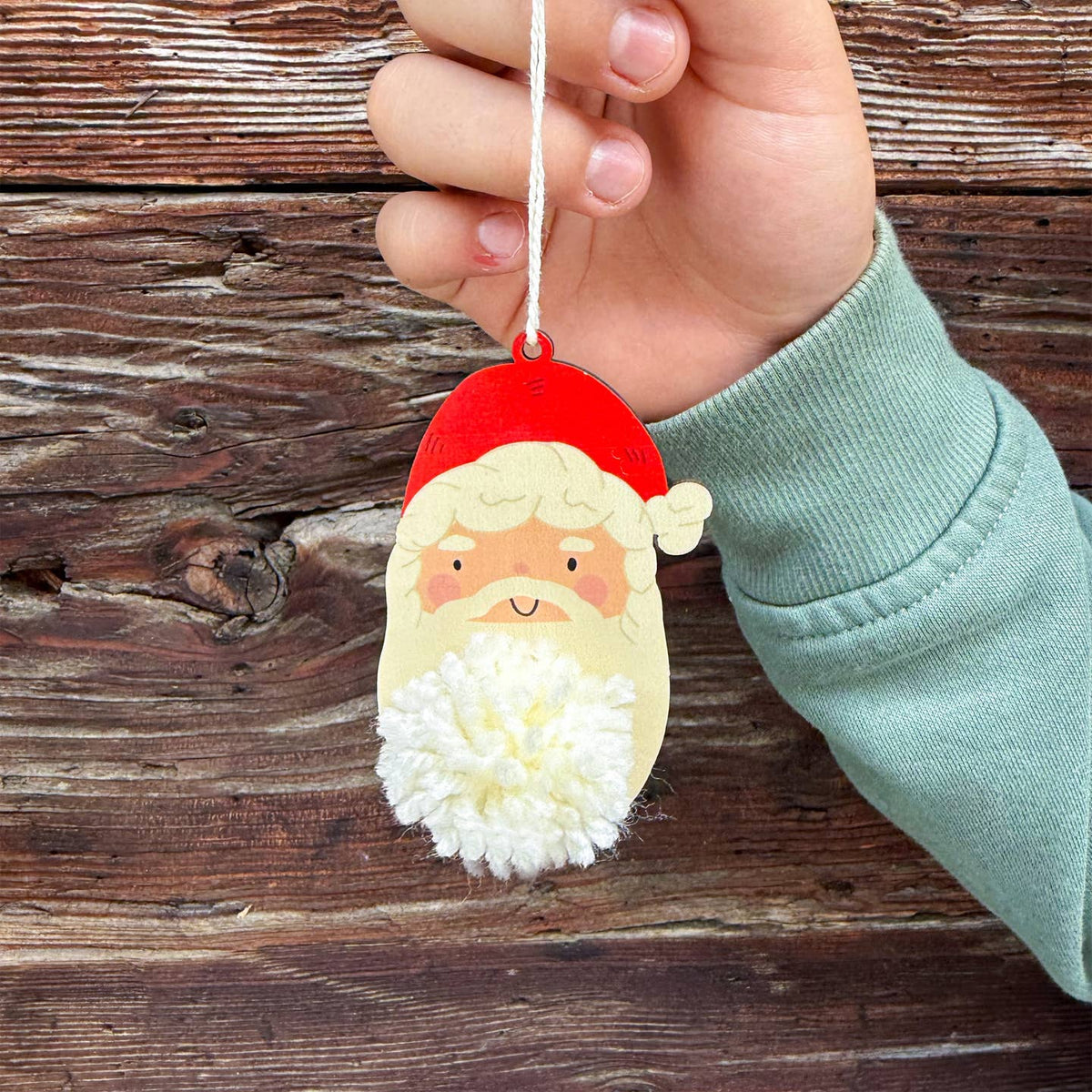 A hand holding a wooden Santa Claus hanging ornament with a white pom-pom beard and red hat, set against a rustic dark wood plank background.