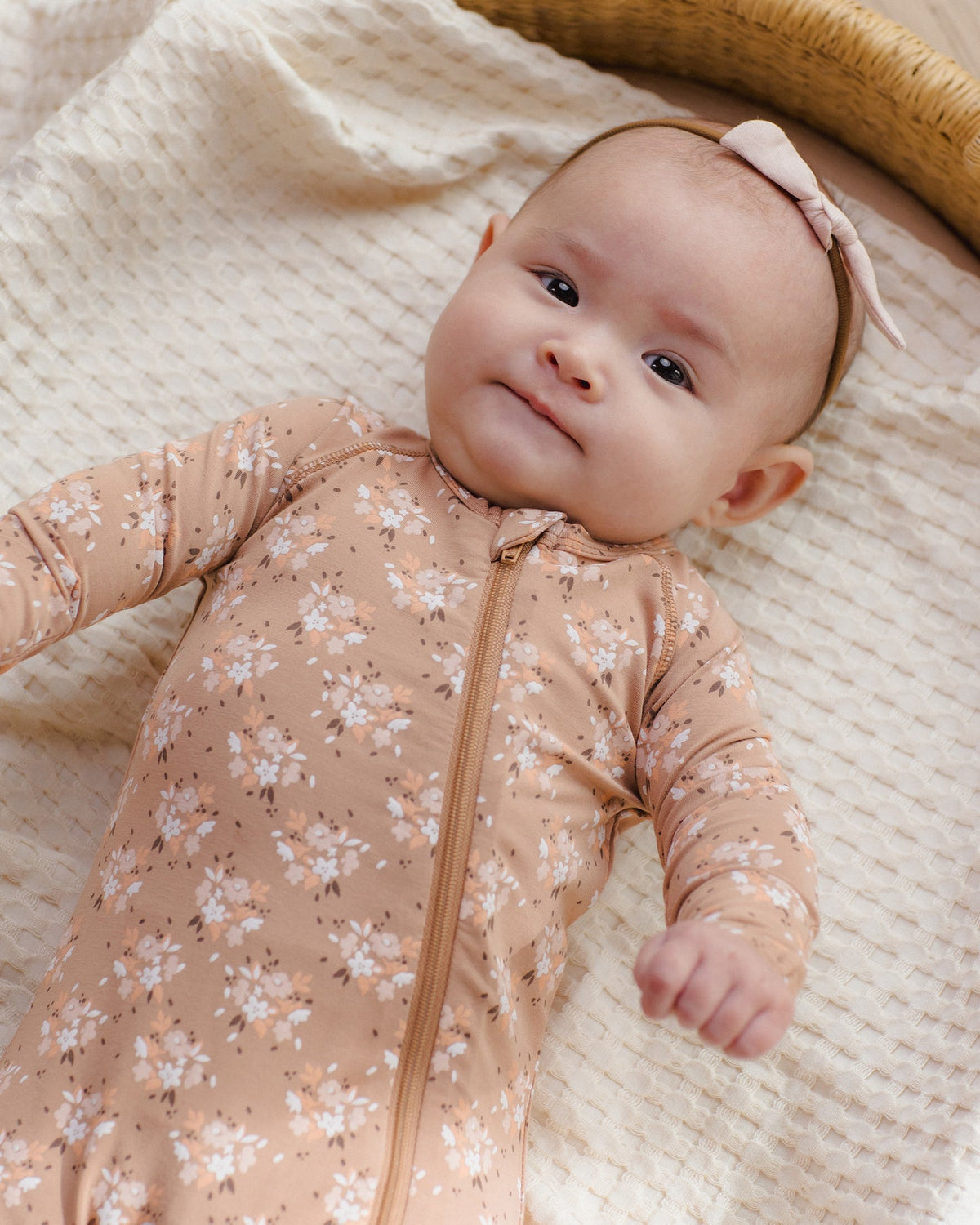 A baby lying on a textured cream blanket, wearing a long-sleeve beige floral zip-up sleeper and a matching pink bow headband.