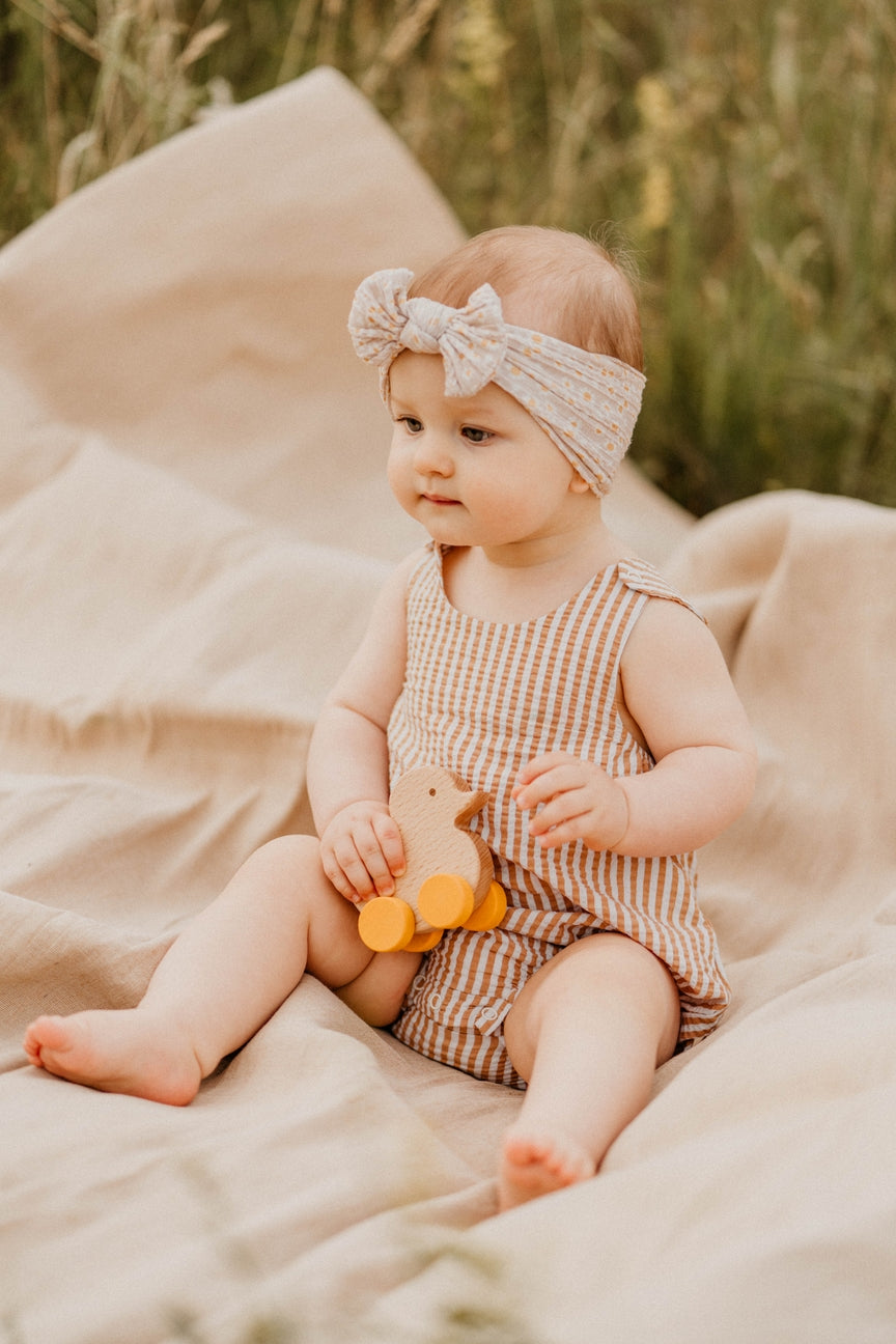 Baby in a checkered romper with a bow headband sitting on a blanket outdoors.