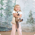 A young girl with blonde hair and a white top smiling while hugging a plush Raccoon by Warmies against a painted winter woodland backdrop.