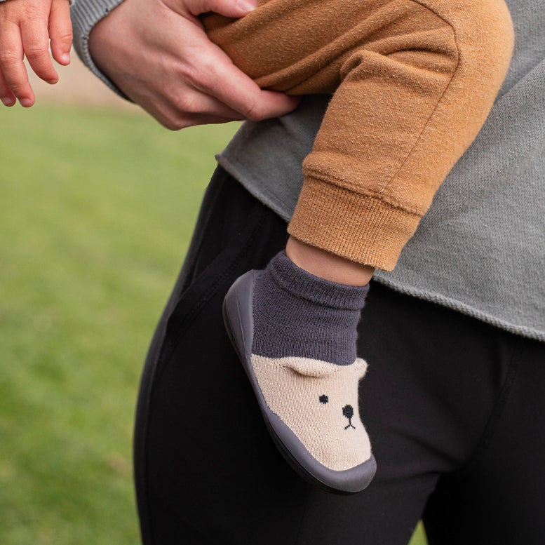 Child's foot wearing a gray sock with a bear design, held by an adult against a grassy background.
