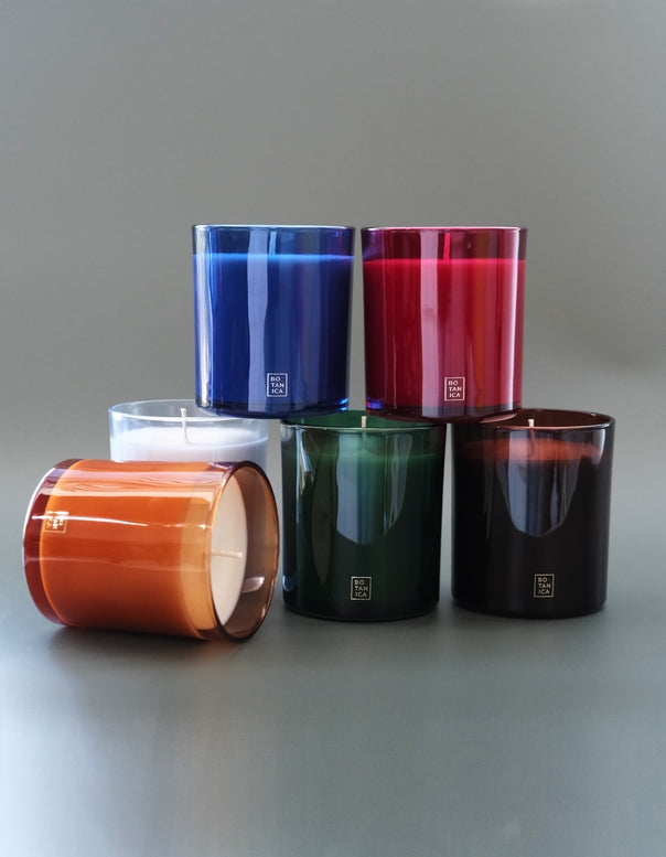 Six glass jar candles in assorted colors, including blue, red, orange, green, black, and white, arranged on a neutral gray surface.