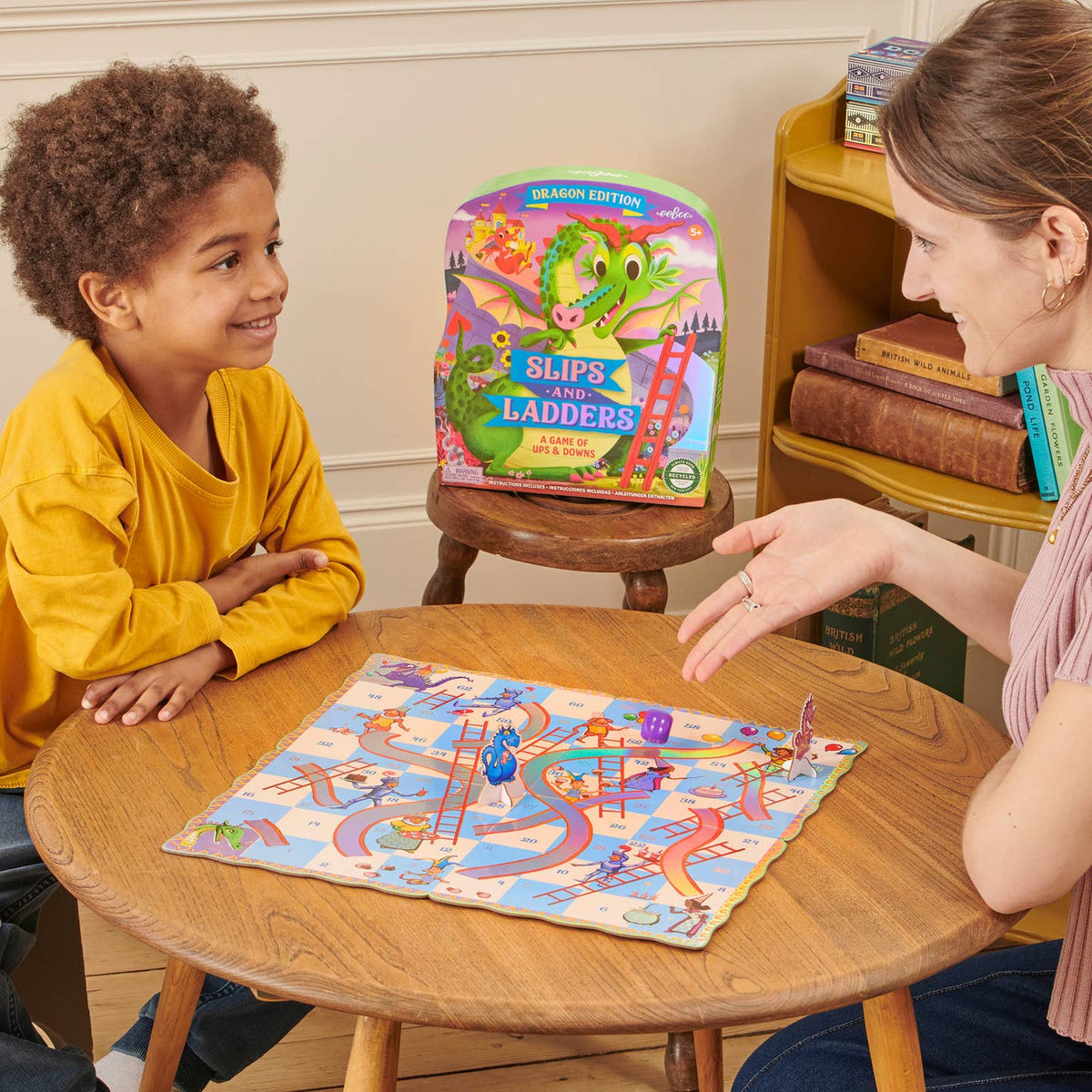 Woman and child playing 'Slips and Ladders' board game in a cozy room.