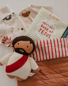 Plush toy of a child dressed as Jesus with a book titled 'Baby's First Book of Jesus' on a bed.