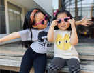 Two young girls sitting on a wooden bench wearing sunglasses and casual clothing.