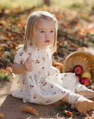 A young toddler girl with blonde hair sits on the ground outdoors among fallen leaves, wearing a cream-colored dress with a small red and brown floral pattern. A woven basket containing red and yellow apples sits next to her.
