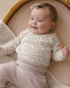 A baby lying on a woven bassinet wearing a cream floral long-sleeve wrap bodysuit with matching pants and a light gray bow headband.