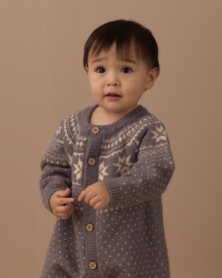 A baby wearing a gray knit cardigan and matching knit pants with cream-colored snowflake patterns.