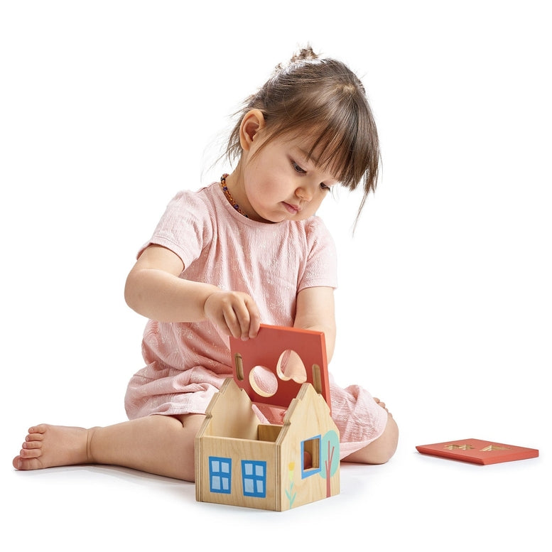 A toddler sits on a white background, gently placing a red wooden shape into the matching slot on the roof of a wooden house shape sorter. Other colorful wooden shapes rest on the surface nearby.