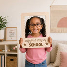 Young girl holding a 'my first day of school' sign in a home setting