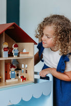 A young child with curly brown hair looking at an open-faced wooden dollhouse by Mentari, arranged on a white table with miniature wooden furniture and small figurines.