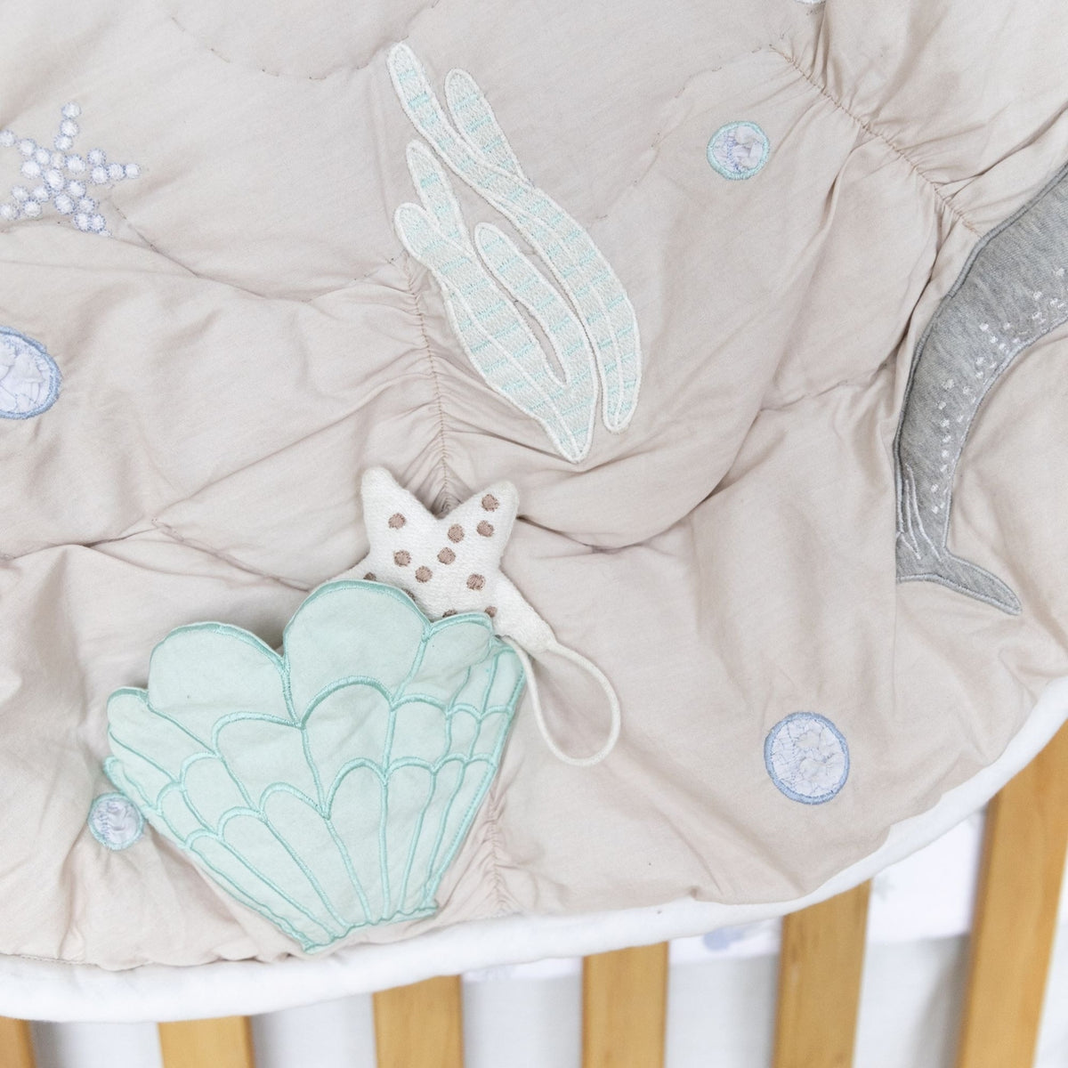 A close-up of a beige quilted baby blanket featuring embroidered sea-themed motifs including a mint green scallop shell, pale green seaweed, a gray crescent moon, and blue coin details, styled on a wooden crib.