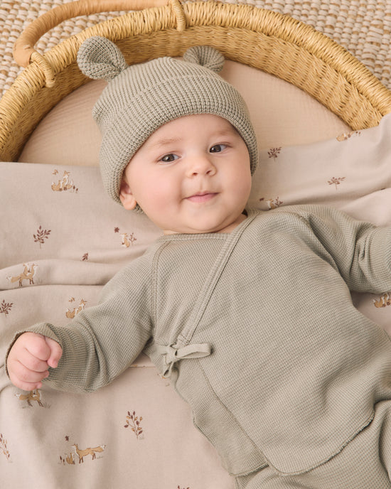 A baby lying on a woven mat inside a basket, wearing a sage green knit bonnet with two small pom-poms and a matching sage green long-sleeve romper. The baby is resting on a beige blanket with a small deer pattern.