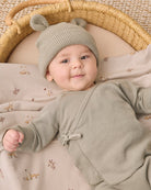 A baby lying on a woven mat inside a basket, wearing a sage green knit bonnet with two small pom-poms and a matching sage green long-sleeve romper. The baby is resting on a beige blanket with a small deer pattern.