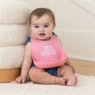 Baby wearing a pink bib with 'Mini Swifthe' printed on it, sitting on a white blanket.