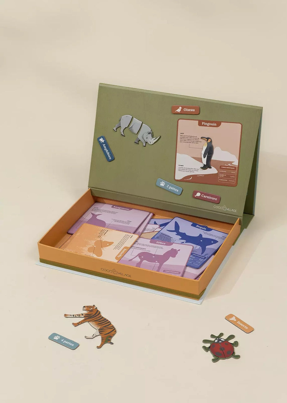 An open educational game box labeled 'PENGUIN CLUB' on a warm beige background. The olive green inner lid features illustrations of a rhinoceros and a penguin, while the open orange-brown base reveals purple and peach-colored cards.