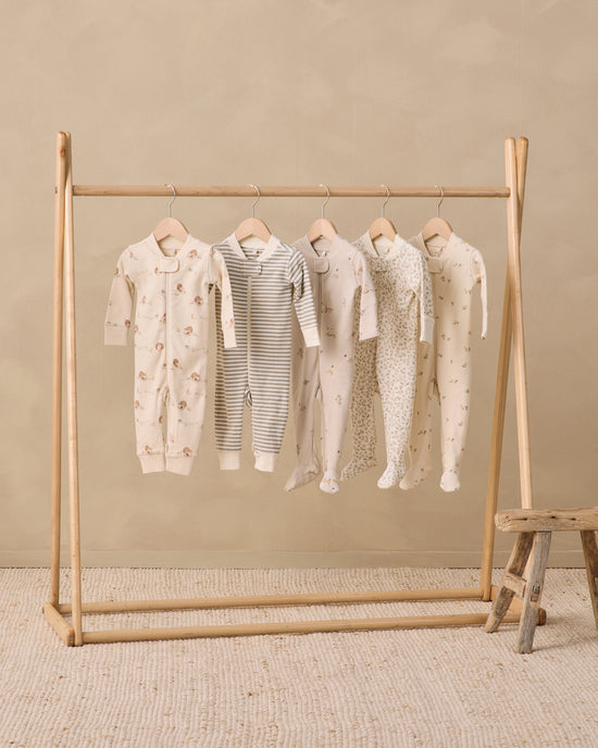 Five baby footed onesies in neutral tones hang on a wooden clothing rack against a beige wall and textured rug. The onesies include cream, gray-and-white stripes, and light floral and polka dot patterns.