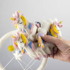 A hand holds a cluster of white felted wool unicorn toys with pastel pink, yellow, and blue accents, attached to a white circular hanging frame against a light gray background.