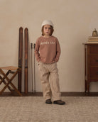 A child stands in a styled indoor room wearing a dusty rose top and matching beige pinstriped pants. , with a cream knit beanie and dark slip-on shoes
