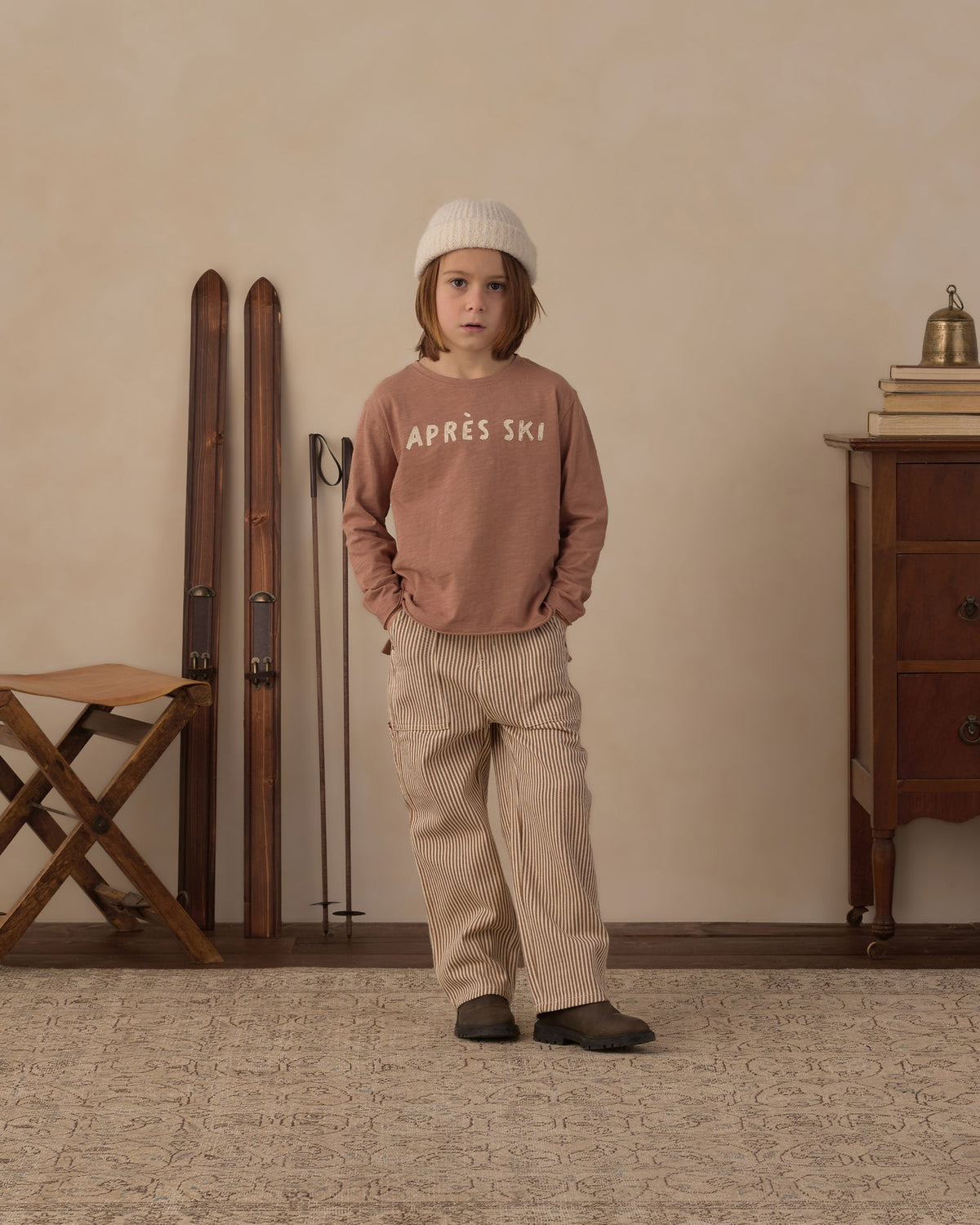 A child stands in a styled indoor room wearing a dusty rose top and matching beige pinstriped pants. , with a cream knit beanie and dark slip-on shoes