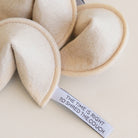 Close-up of a plush fabric comforter or cushion in a warm cream/beige color, with folded stitched edges and a small sewn label reading “THE TIME IS RIGHT TO SHRED THE COUCH.”.