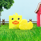 Yellow chick toy with packaging on grass in front of a red barn and green trees