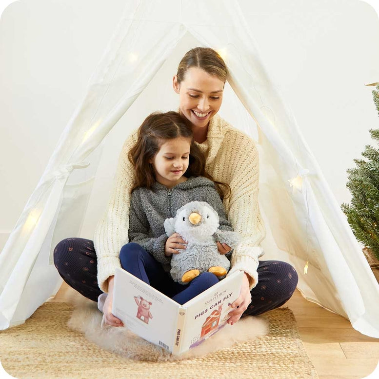 A woman and a young girl sitting inside a cozy fabric tent, reading a picture book together while holding a gray penguin plush toy.