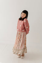 A young girl with black hair is smiling and looking to the side while wearing a pink textured knit cardigan with white pom-pom details and a long pink patterned skirt with a floral lace hem, paired with beige shoes, against a bright white studio background.