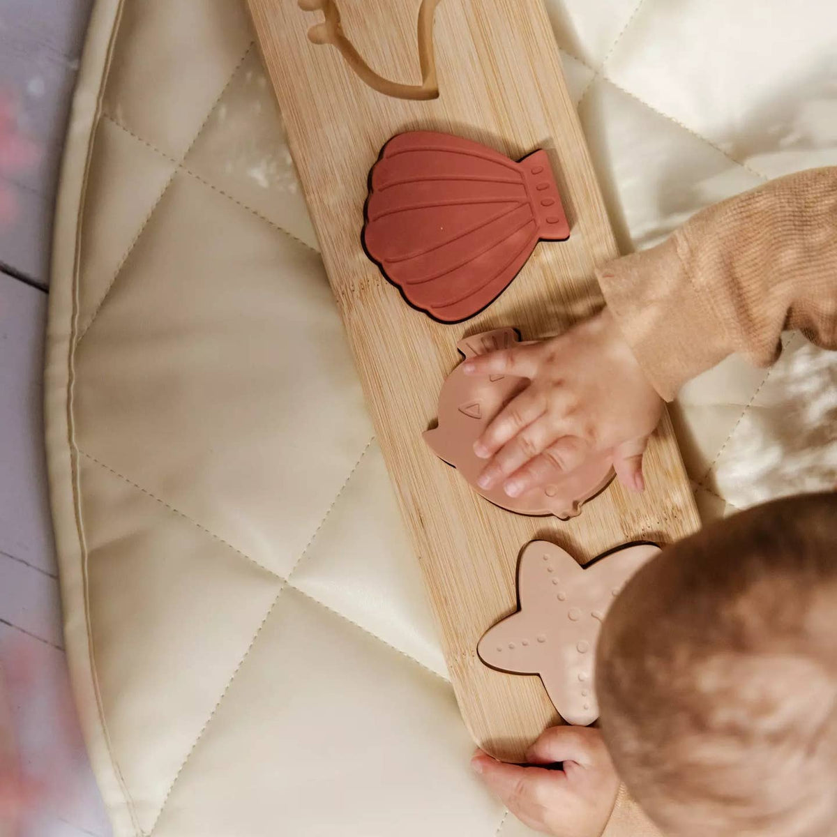 A baby's hands and head are visible as they explore a wooden sensory puzzle board with shell, cat, and star shapes on a quilted cream play mat.