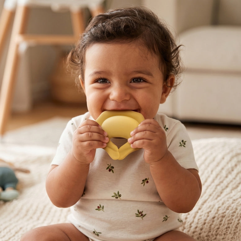 A baby wearing a white short-sleeve bodysuit with small green leaf prints is sitting on a textured rug and biting a yellow teething toy.