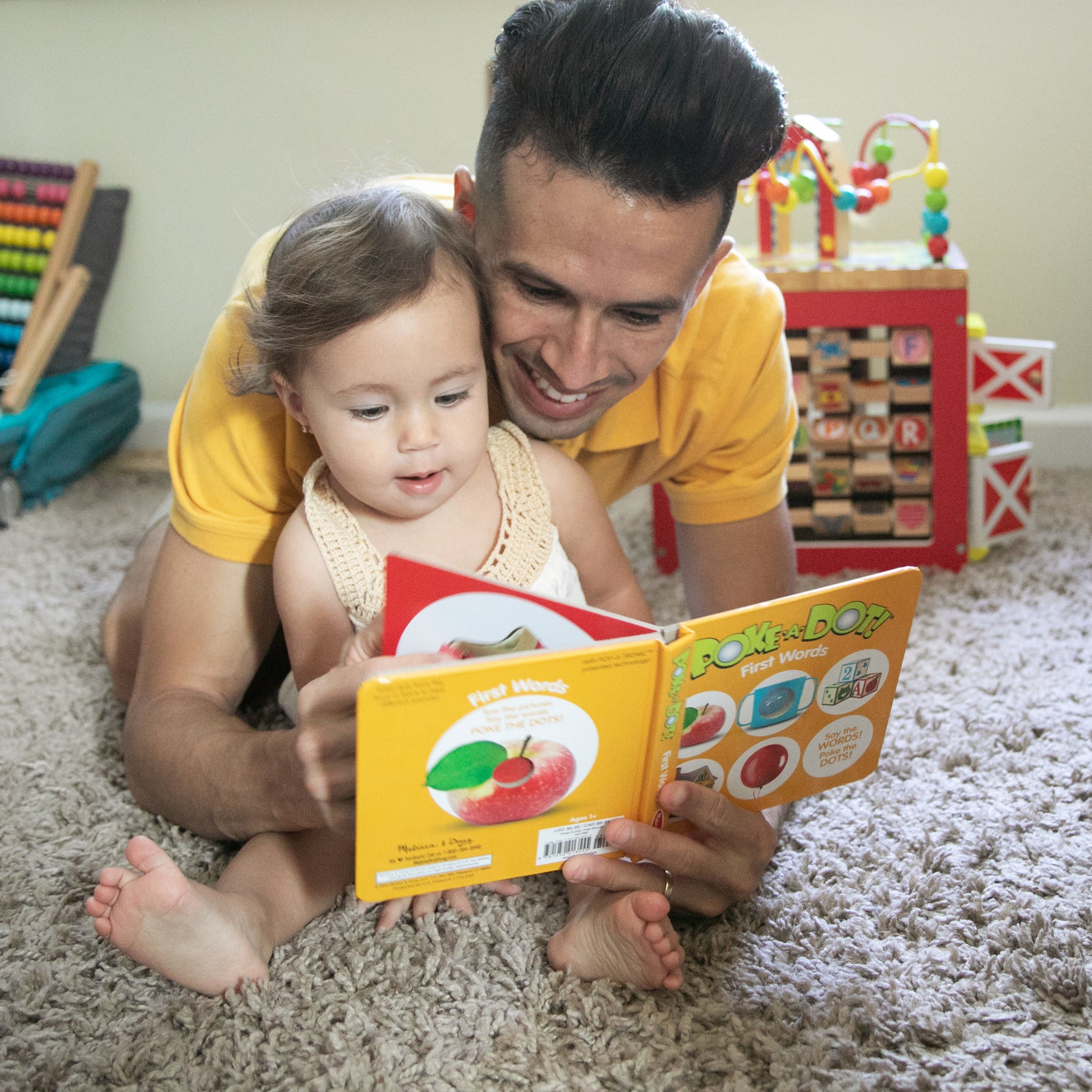 A toddler and a man sitting together on a carpeted floor, looking at an open interactive board book titled Poke-a-Dot! First Words.